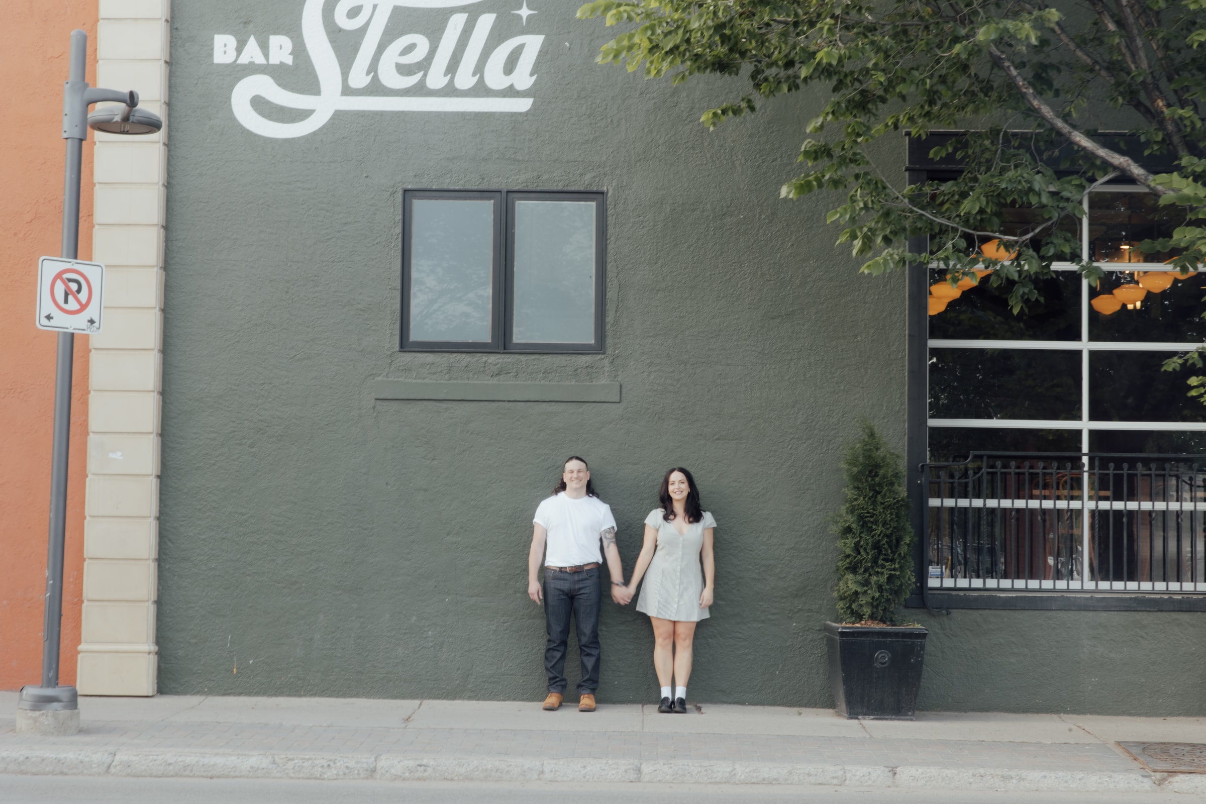 Briana and Ethan standing hand in hand against a deep green wall under a hand-lettered Bar Stella sign.