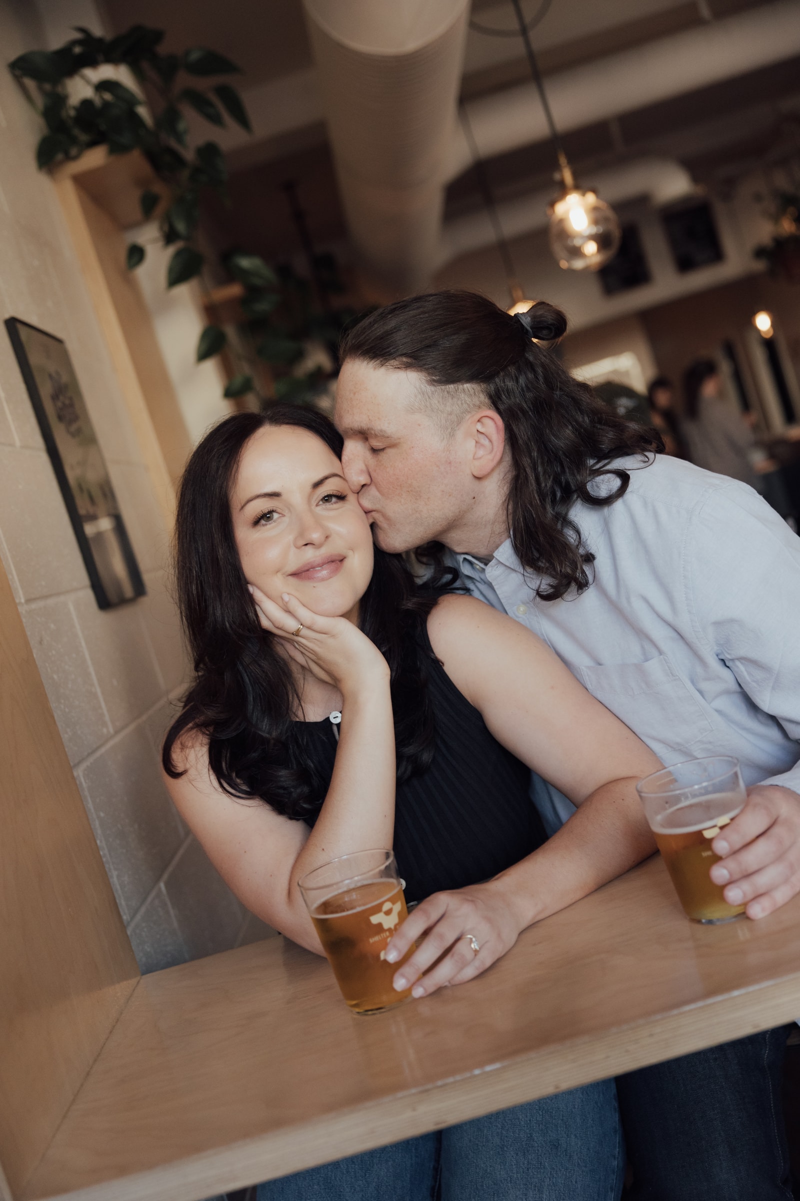 Ethan kissing Briana on the cheek at a taproom table, both holding glasses of beer as she smiles at the camera.